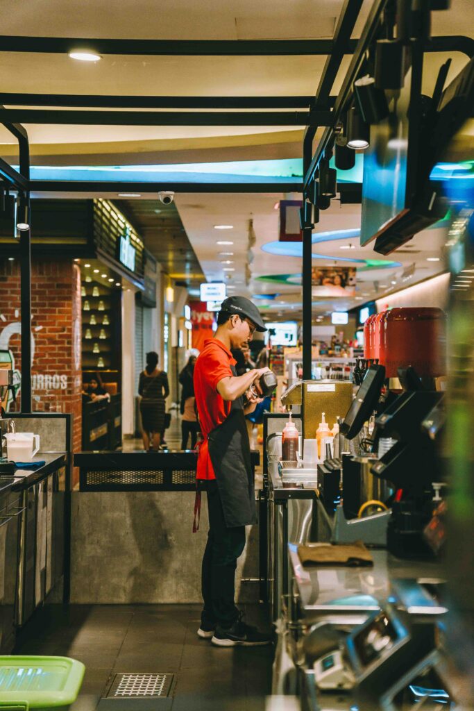 A worker in a Kuala Lumpur mall fast-food shop serves customers amid bustling activity.